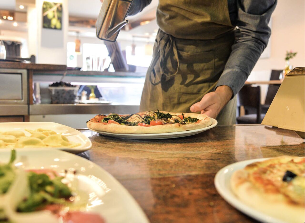 Kellner gibt Olivenöl über eine vegetarische Pizza, daneben Ravioli und Carpaccio auf der Theke