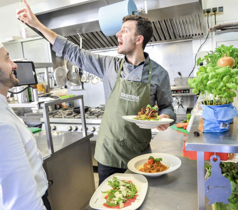 Waiter in a green taormina apron carrying pasta and carpaccio plates in the open kitchen