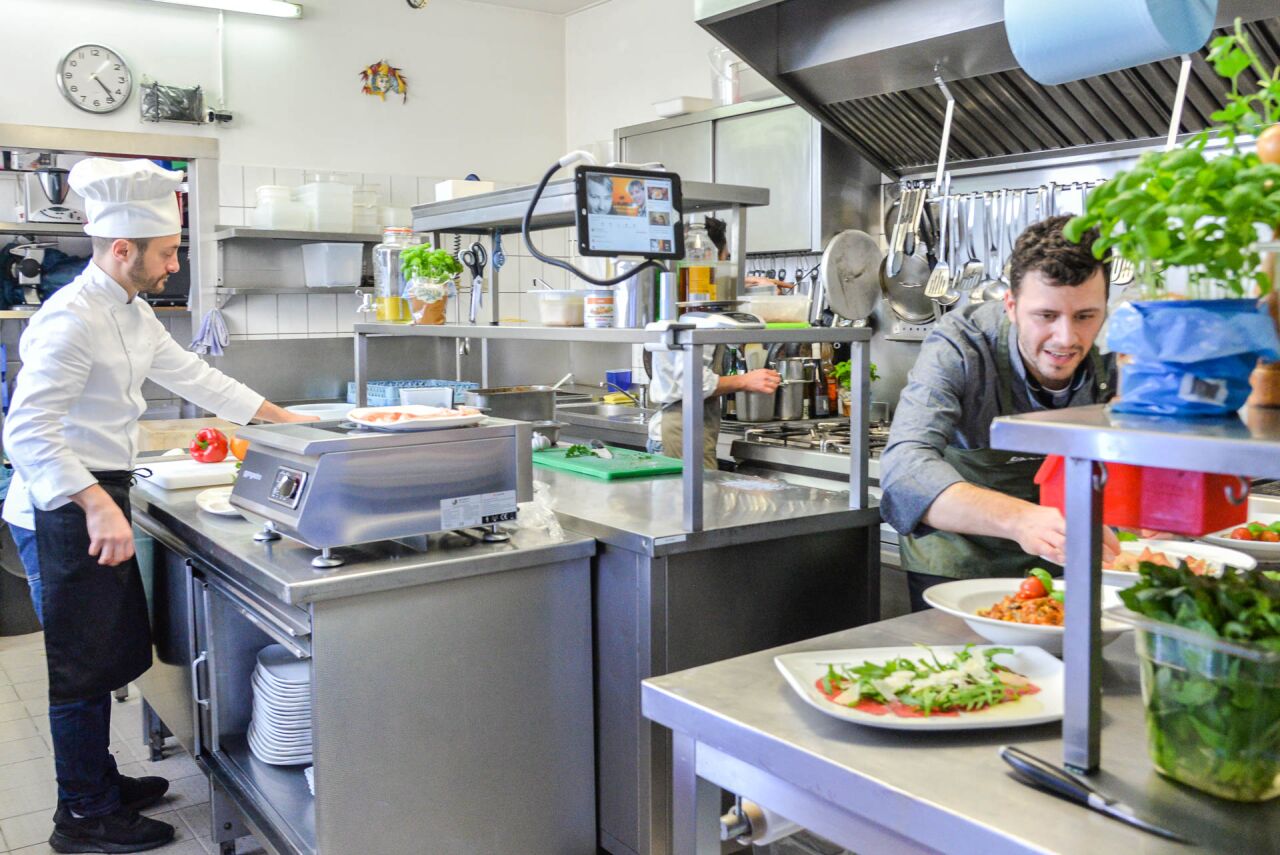 Open kitchen: chef in whites plating rocket carpaccio at the stainless steel counter, kitchen helper behind