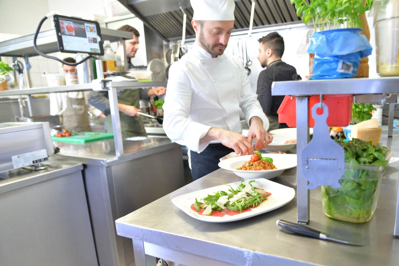 Koch richtet in der Küche Pasta mit Tomatensauce und Rucola-Carpaccio an, im Hintergrund das Küchenteam