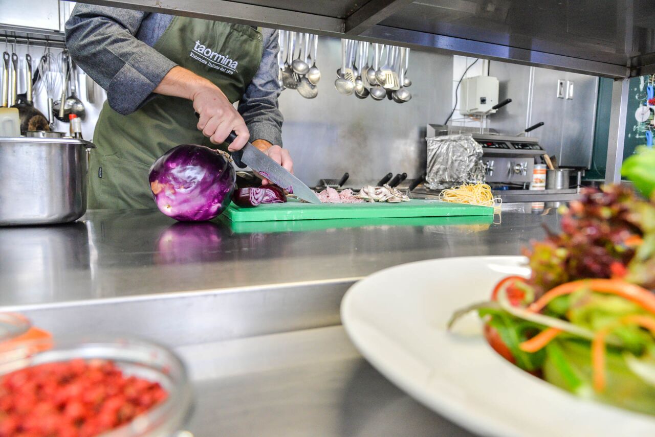 Koch in grüner taormina-Schürze schneidet eine Aubergine, vorn ein Salatteller