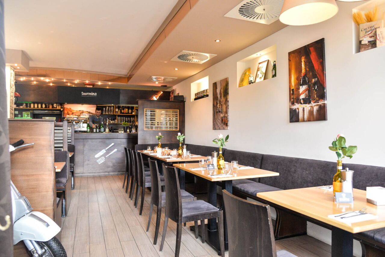 Langer Gastraum mit Sitzbank, gedeckten Holztischen, Vespa und Bar im Hintergrund