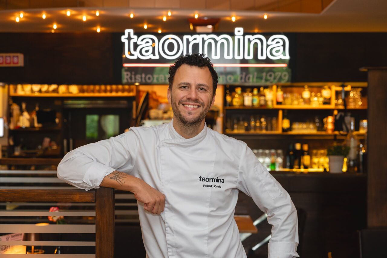 Fabrizio Costa in weißer Kochjacke lehnt lächelnd an einem Geländer, dahinter das leuchtende taormina-Schild und die Bar des Restaurants